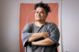 Pacific art. Image is of a person with dark curly short hair, folded arms and grey shirt looking askance at the camera and standing in front of an ochre coloured wall hanging.