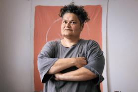 Pacific art. Image is of a person with dark curly short hair, folded arms and grey shirt looking askance at the camera and standing in front of an ochre coloured wall hanging.