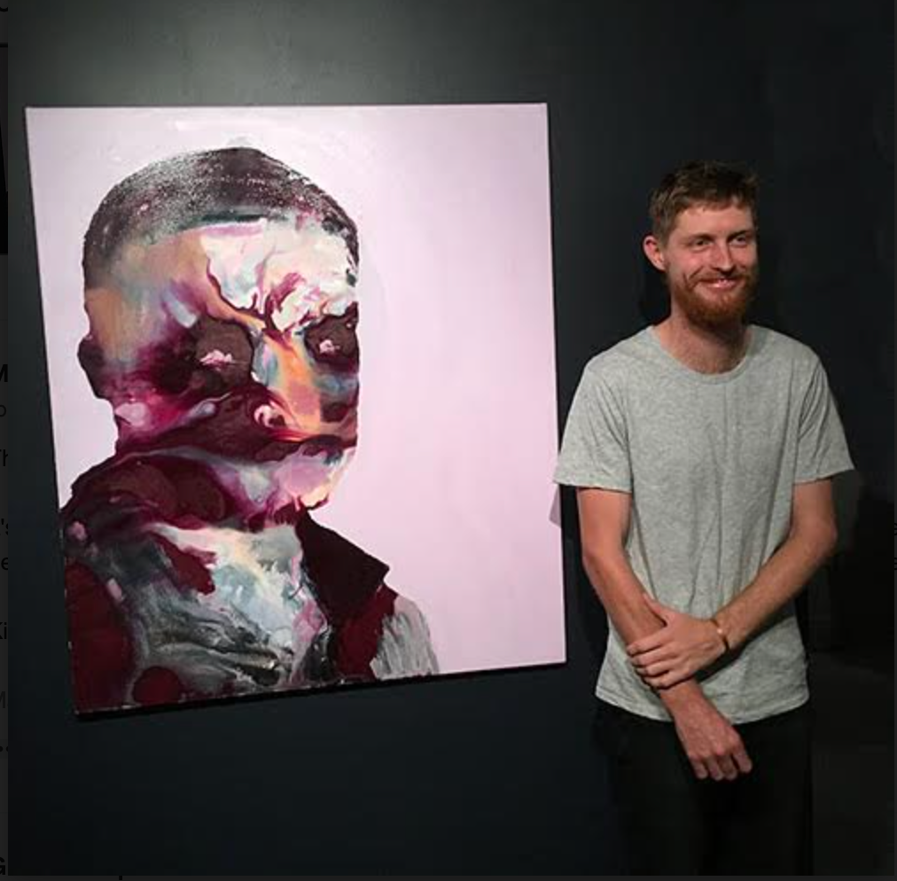 other work. man with beard and grey t-shirt standing in front of colourful abstract painting of a face.
