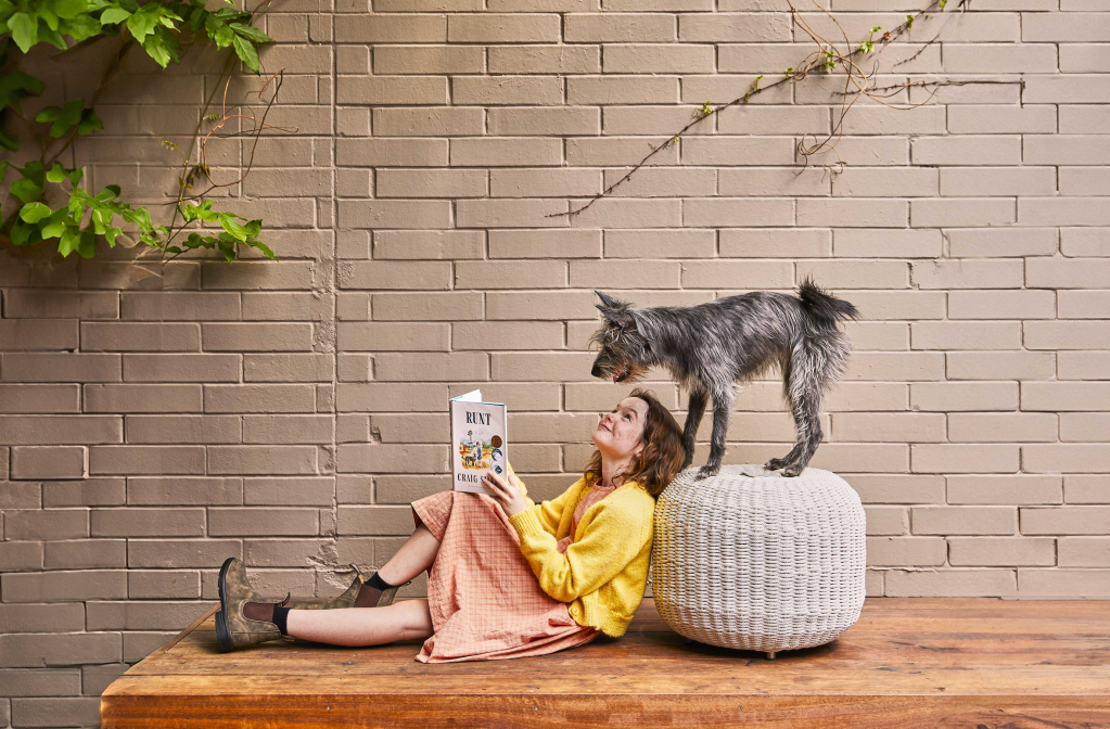 Runt. Image is a young girl lying on a deck against an ottoman, with a book in her hands, but looking up at a grey haired dog above her standing on the ottoman. She is wearing a yellow top, pink dress and brown Blundstone boots.