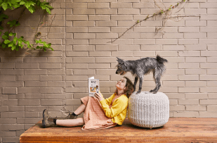 Runt. Image is a young girl lying on a deck against an ottoman, with a book in her hands, but looking up at a grey haired dog above her standing on the ottoman. She is wearing a yellow top, pink dress and brown Blundstone boots.