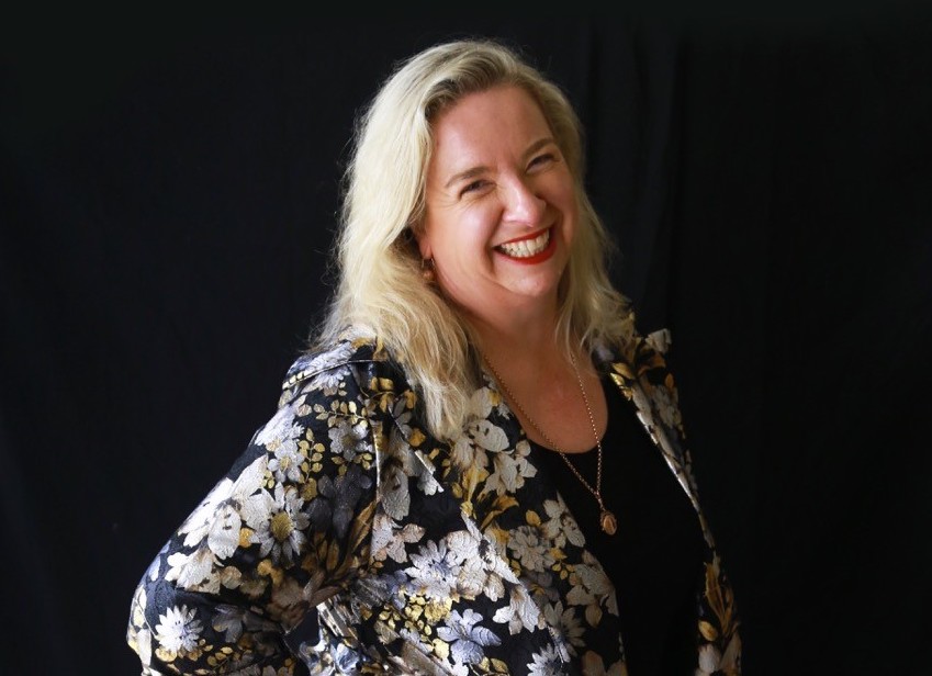 Muddiman. Laughing woman with blonde hair and wearing a gold and black top.