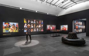 Tarnanthi. Image is gallery space full of colourful Namatjira paintings and a full-size sculpture of a First Peoples bearded man in a white cowboy hat.
