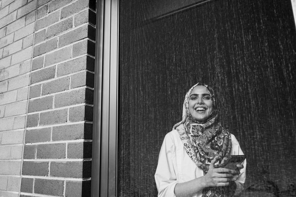Wahija Pervez. A black and white image of a young woman in a headscarf, laughing and holding a mobile phone. A brick wall frames her on the left. She is a fashion lecturer and PhD student.