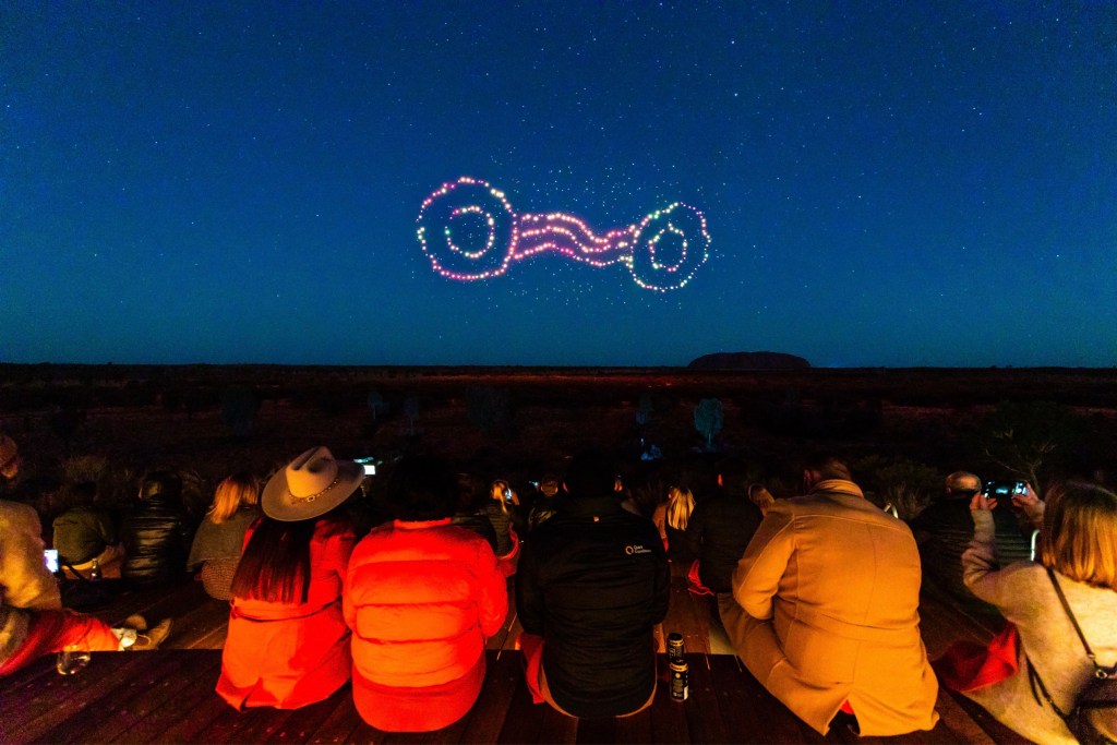 People sitting in outdoors watching drone show in the sky over the ancient site Uluru