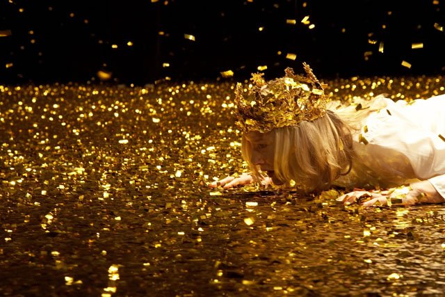 Shakespeare. Image is top half of a woman lying flat on stage covered in gold with flakes descending. She is in a white costume, wearing a gold crown with her hands to the side of her face flat on the groun and her chin raised up to look forward.