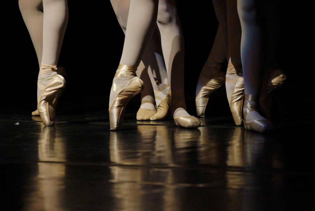 regional ballet. Photo of ballet dancers' lower legs wearing ballet shoes on a black stage.