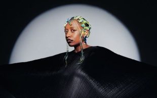 Salvant. Image is of a spotlight woman wearing a black cloak with arms outstretched to the side to fill half the spotlit space, looking at the camera and with multicoloured hair.