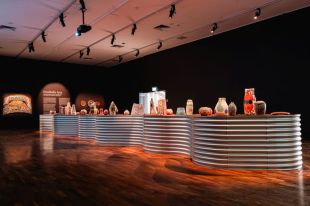 Ernabella Arts. Warka Wiru. Image is a gallery space with a corrugated snake like plinth on a polished wooden floor bearing a wide selection of diverse ceramics.