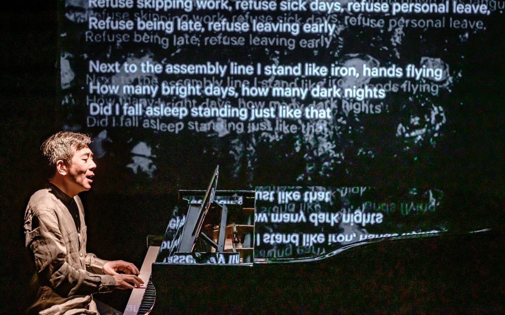 I Swallowed a Moon Made of Iron. Image is a man sitting at a grand piano playing and singing, while on a screen behind him are translations of Chinese poetry into English.