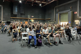 Hall of people on chairs voting with arms raised.