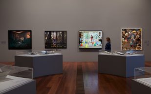 ‘Photography: Real and Imagined’ Image is a grey walled gallery with one woman looking at four photographs on the back wall and grey plinths under glass showcases in the foreground with more work on them.
