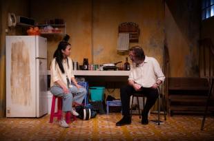 Flake. Image is a grungy kitchen theatre set with two people sitting at a table, a young woman of Asian appearance on a stool with white shirt, grey trousers and hands on knees and an older bent over man with black trousers, white shirt, glasses and straggly grey hair.