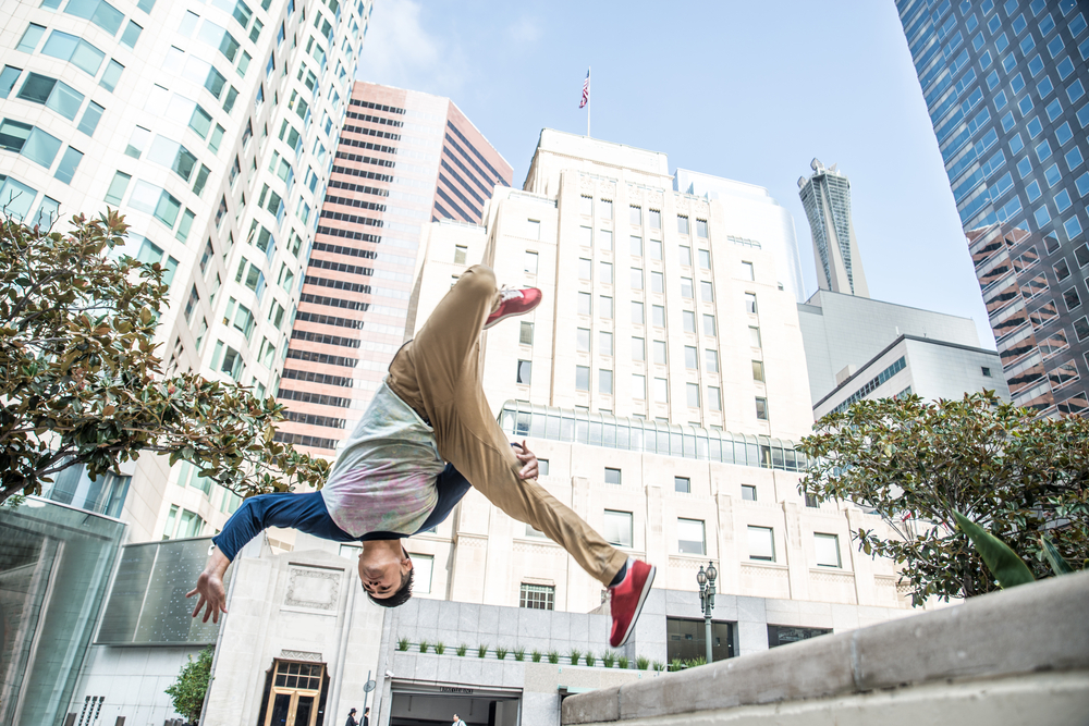 appointments. Image is a parkour performer caught mid-backflip on a city street. Tall buildings rise behind him, his head is pointed towards the ground and his arms and legs are outstretched mid-flip.