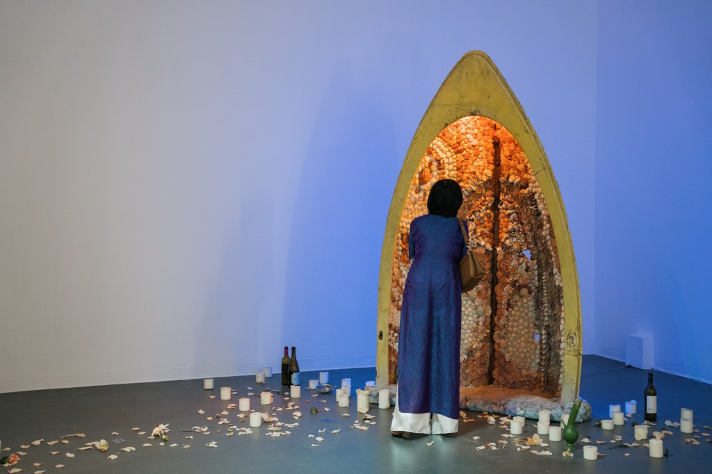 Studios: 2023. Image is a woman in long blue dress and with a brown shoulder bag, with her back to camera looking at an art piece made of half a rowboat standing on end, full of shells and other additions, with candles, empty bottles and a bong on the floor around it.