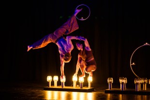 Circus. Image is two acrobats, one doing a one-handed handstand on a row of footlights and the other crouching and helping the first to balance.