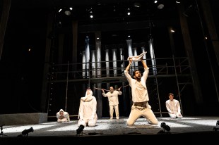 This Rough Magic. Image is a bare stage with several actors, kneeling, sitting or standing, one holding up a stick, and another holding a cloth that could symbolise a child.