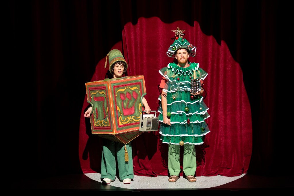 A Very Jewish Christmas Carol. Image is two people standing on stage in a spotlight in front of a red curtain. One is dressed as a present, the other as a Christmas tree.