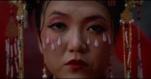 The Bridal Lament. Image is a close-up of an Asian woman's face with a complex headdress, heavy makeup and facial adornments in the form of eight white pearl tears. She looks sad.