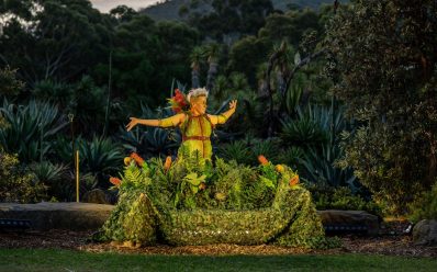 'Shakespeare in the Garden – A Midsummer Night’s Dream'. Image is a green leafed clad fairy atop a green leafy piece of set with a background of trees.