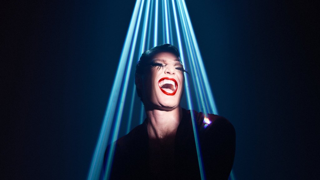 Hobart Festival of Comedy. Image is of a man wearing a lot of makeup and sporting a huge open mouthed grin, while standing in a blue strobing spotlight.