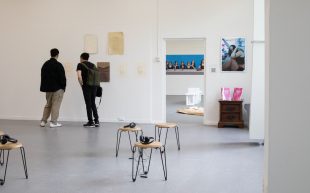 Graduate. Image is of a couple of gallery visitors standing by the far wall of a gallery looking at art, with other pieces in the foreground.