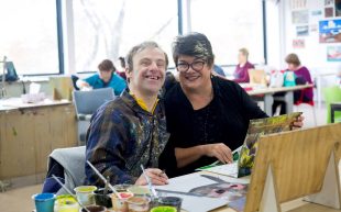 Sue Roff. Image is an artist in a black T shirt, at work and smiling at the camera, next to a woman with short grey hair and glasses who is also smiling at the camera.