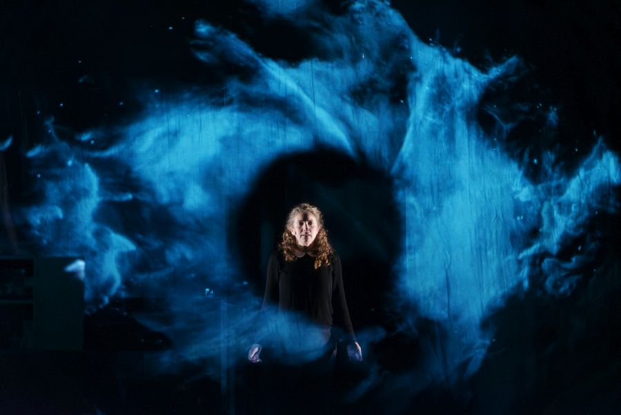 At the End of the Land. Image is a swirling blue haze with a black circle in the middle in which stands a woman with black top and shoulder length hair.