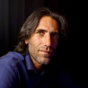 refugee. Image is head and shoulders of man of Middle Eastern appearance with shoulder length dark hair and blue shirt against a black background. He is smiling slightly.