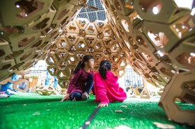 Education. Young girl in pink dress sits inside hive-like strucure on green grass as part of art for kids.