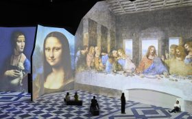 Leonardo da Vinci – 500 Years of Genius. Image is massive gallery space with scattered viewers and a huge all taken up with a Renaissance painting of Christ and the last Supper and another of the Mona Lisa.