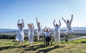 Audio description app to make the arts accessible for blind and low-vision patrons in line for $30,000 entrepreneurial prize. Photo: O&J Wikner Photography. Five figures wearing white in a natural hilly landscape raising their arms in midair.