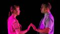 Dead Man's Cell Phone. Image is two actors, one male, one female in a pink light against a black background. They are facing one another and touching their fingertips together.