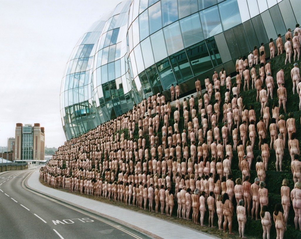 Nude Spencer Tunick. Image is lines upon lines of naked people behind each other on rising levels, standing in front of a curved glass structure.
