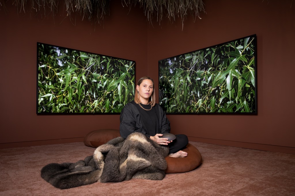 Lucy Simpson. Image is a young woman sitting cross legged in between two screens filled with foliage. There is a possum cloak over her legs.