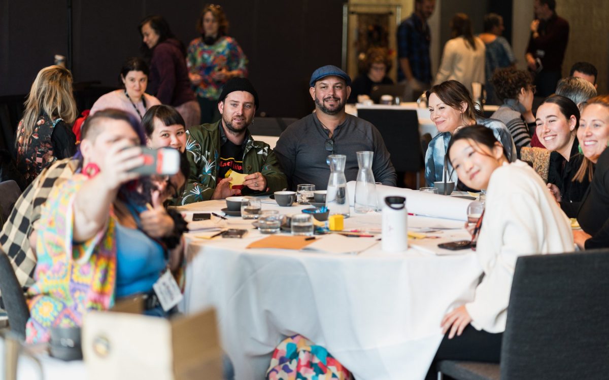 People gathering at Artlands 2023, hosted by Regional Arts Australia. Image is diverse people sitting around a dinner table with a white cloth. One of them is holding up a phone taking a selfie.