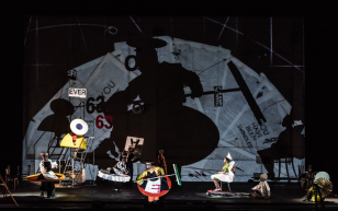 Sibyl. image is a range of performers on a dark stage in wide outlandish costumes, one of whom is holding a large broom and casting a huge shadow on a backdrop that is also covered in projections of newspaper front covers.