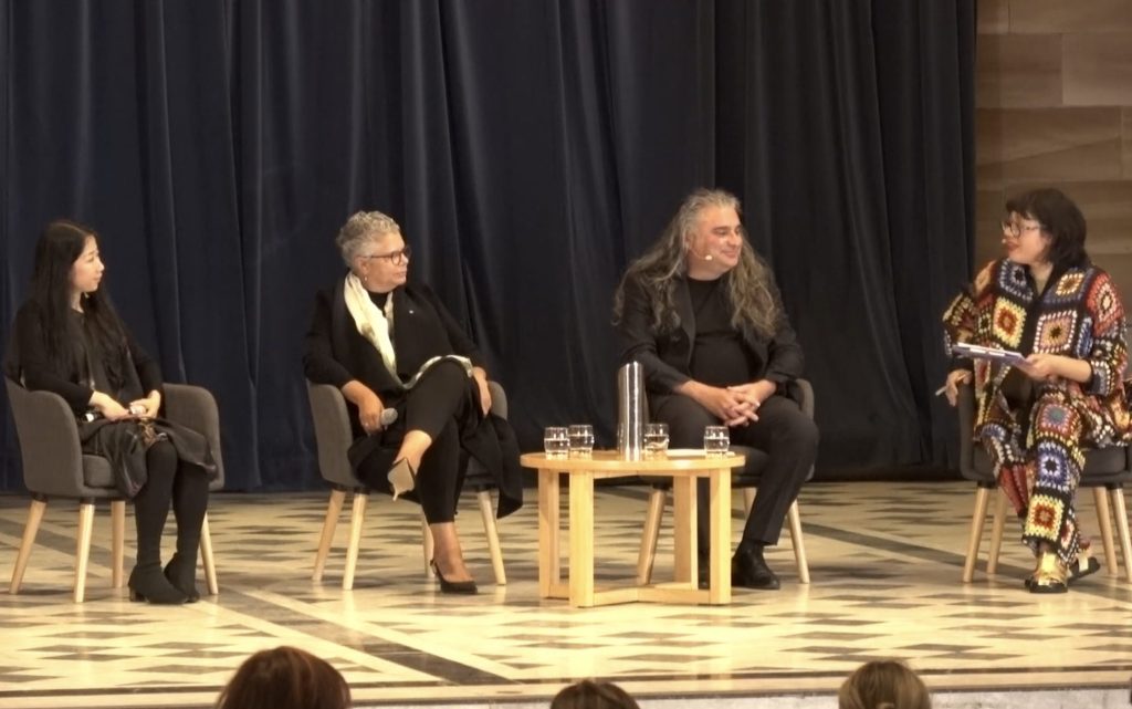 What art ought to be. Image is of four people sitting on a stage in front of an audience, in conversation. On the left a woman of Asian appearance, with long dark hair, on her right a woman of Aboriginal appearance in grey and on her right a Caucasian man in black with long grey hair - and they are all looking at a woman on the far right in colourful crocheted attire.