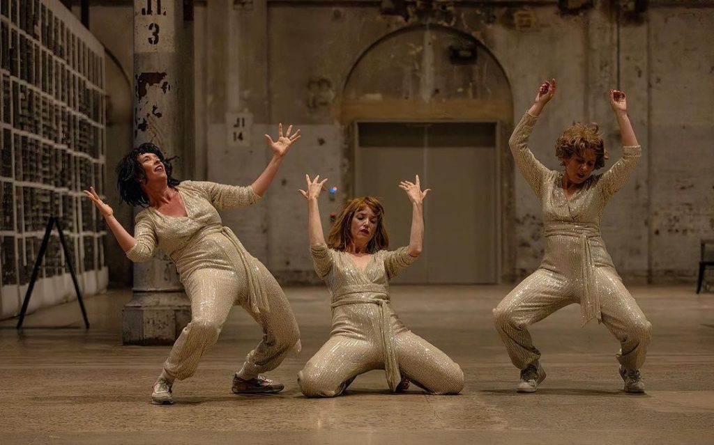 Nighttime Righttime. Image is three women dressed in light gold shimmering one-pieces with sashes, gyrating in a large empty room.