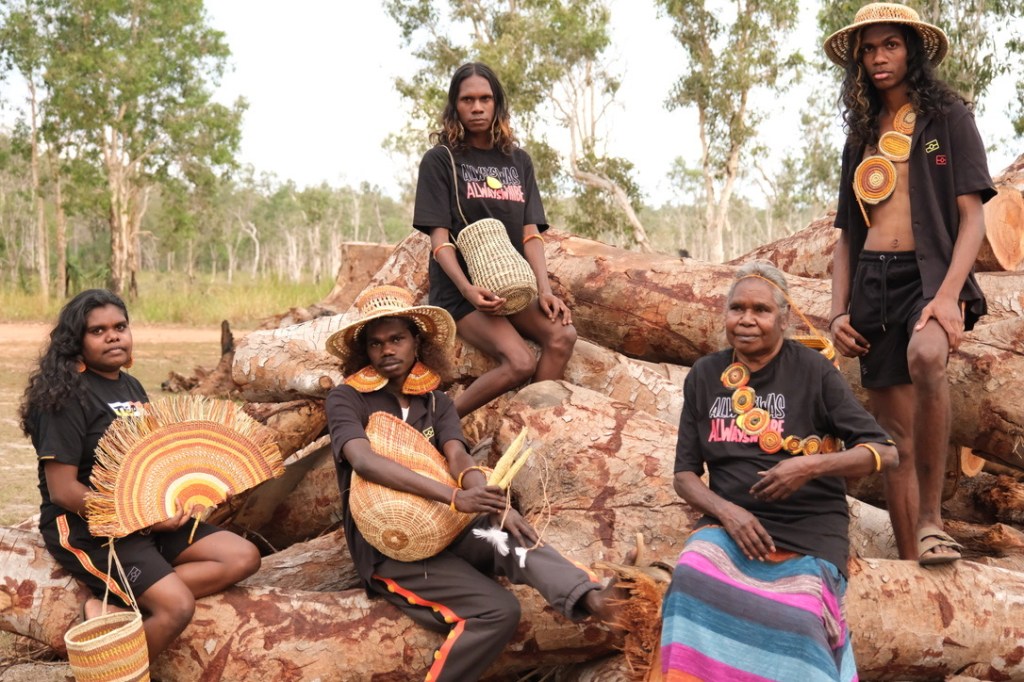 Fashion. Five First Nations people sit on an outcrop of rocks in black T shirts and wearing or displaying a range of handmade fashion accessories.