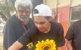 Blood. An Elder Aboriginal man with white hair and beard, stands behind a young Aboriginal man with a backwards white baseball cap and black T shirt with a sunny emblem on the front.