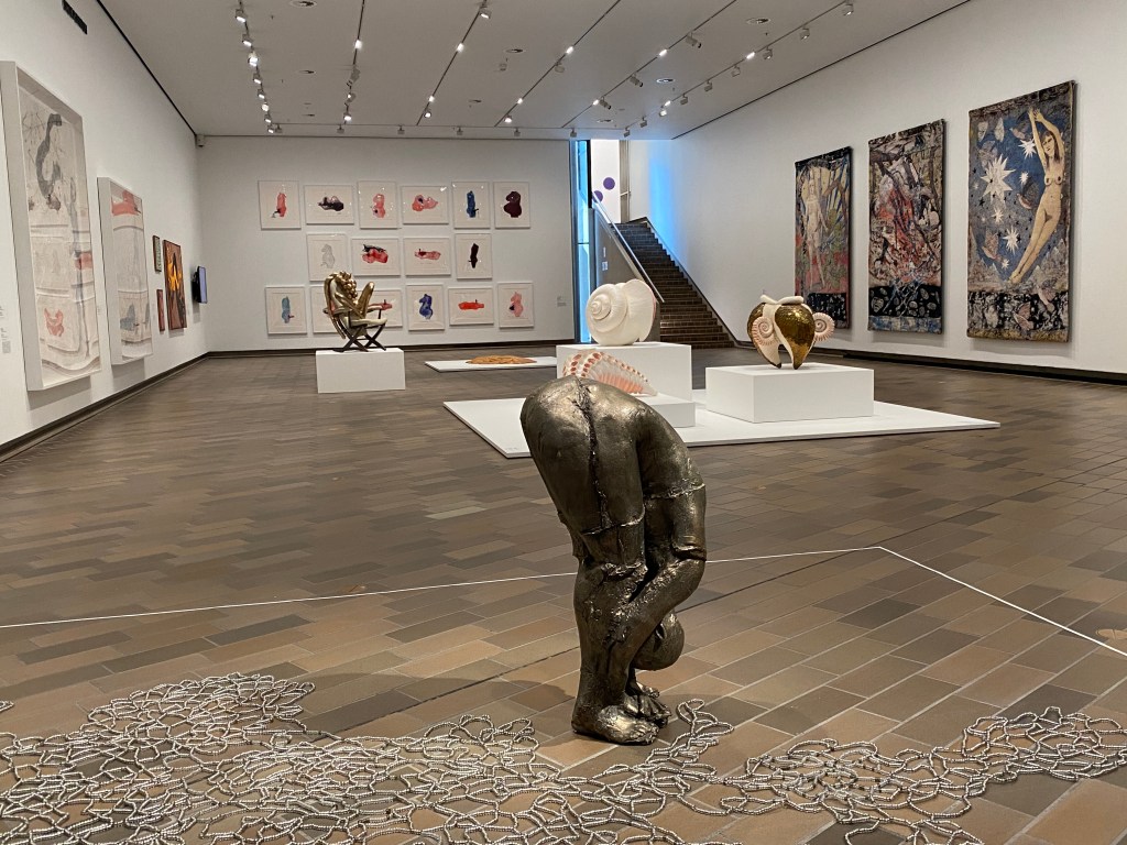 Deep inside my heart. Image is a bronze figure bending right over from the waist and holding their feet. There are beads all around their feet. Elsewhere in the gallery space are plinths with sculptures on them and art works on the walls.