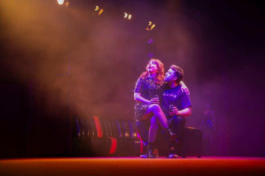 In the Club. Image is a theatre set of a dark smoky club with a black and red striped bench seat and a young man sitting with a young woman on his knee.