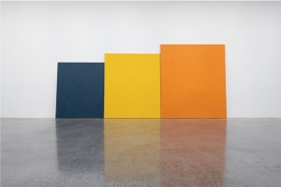 Here and There. Image is of three four-sided mono-coloured images immediately next to each other along a gallery wall. The dark one is the shortest, next to it is a slightly taller yellow one and the tallest orange one is on the far right.