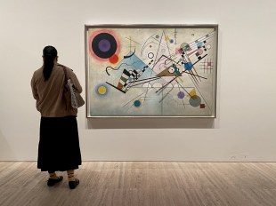 kandinsky. image is the back of a woman in long dark skirt and grey top with bag over her shoulder and a dark ponytail standing to the left of a modernist painting full of squiggles and a large dark sun-like circle in the top left corner.