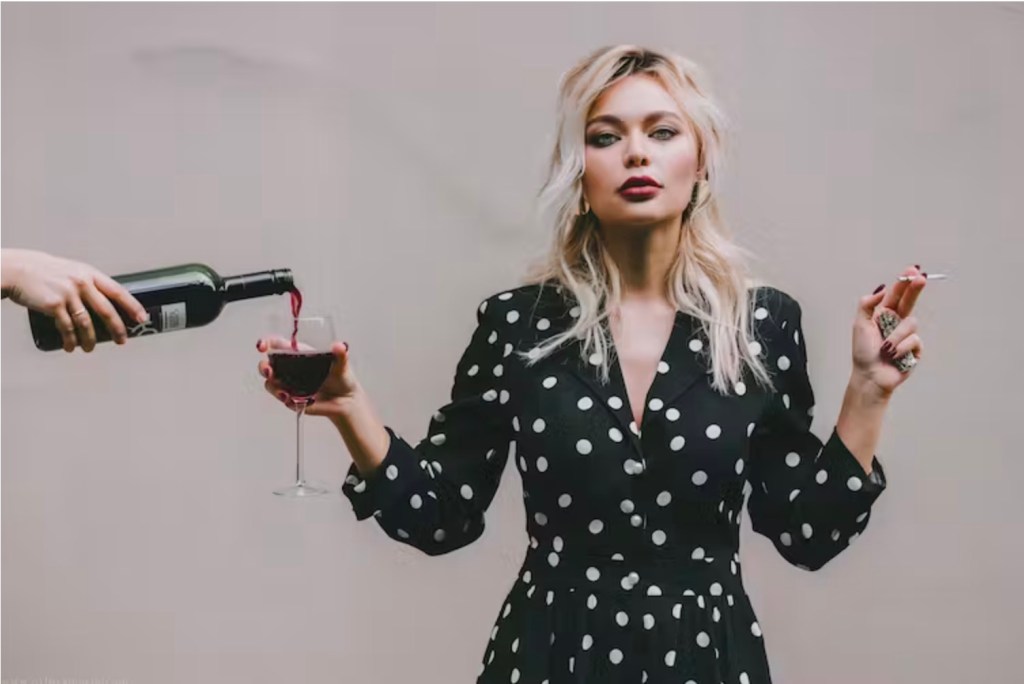 sad bad girls. Image is a blonde woman in green dress with white spots holding out a glass with the hand of someone out of frame pouring wine into it.