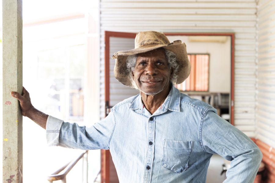 Aboriginal artist Mervyn Street, pictured in 2023, standing on a balcony of an old art shed. He wears a blue shirt and a cowboy hat.