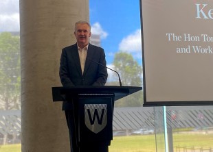 state of the arts. Image is of a man standing in front of a pillar and window next to a screen, and behind a lectern with his hands clasped in front of him. Behind him through the window is grass and a blue sky with some white cloud,