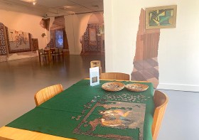 Parlour Parlëur. Image is a golden table with green top, half-completed jigsaw and shallow dishes, with torn images adorning the walls of the gallery space.
