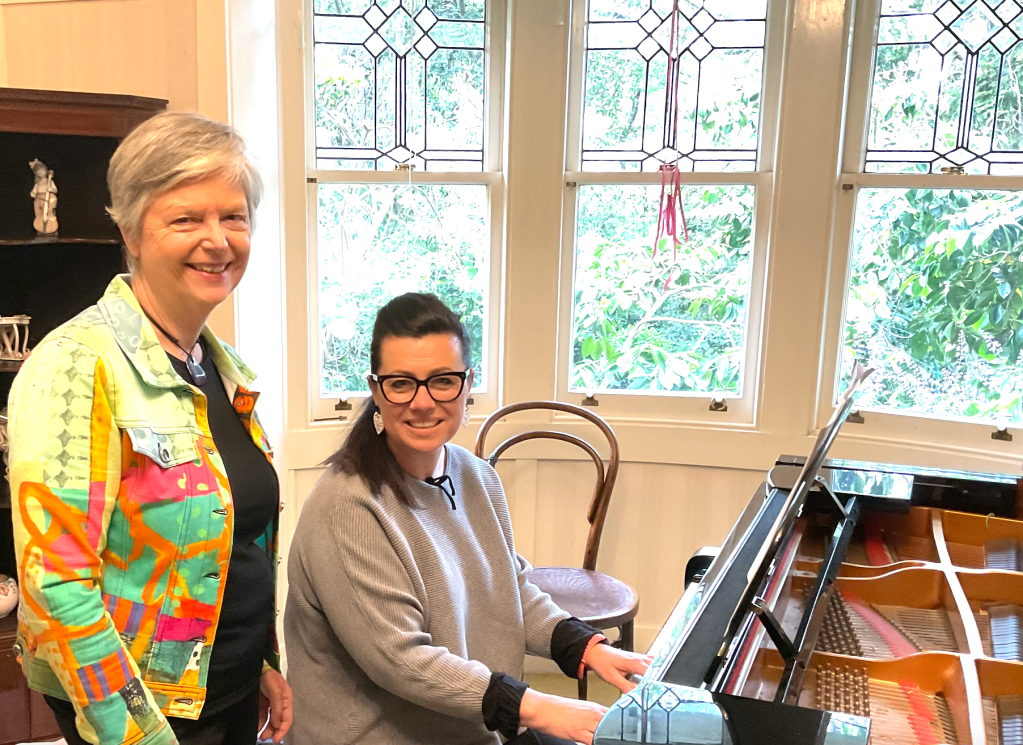 Older white woman with grey hair stands beside younger woman with dark hair and glasses, seated at the piano, mentoring and teaching her.
